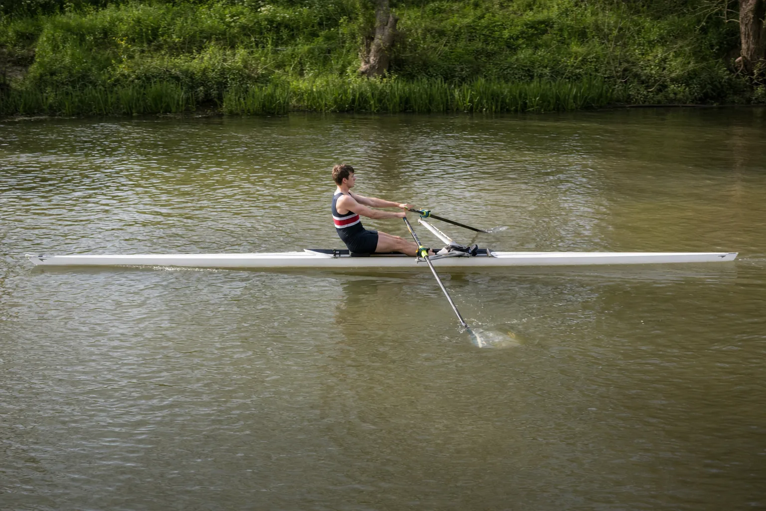 A single sculler on the drive, both blades fully buried, legs pushing down and the boat moving with a bow wave