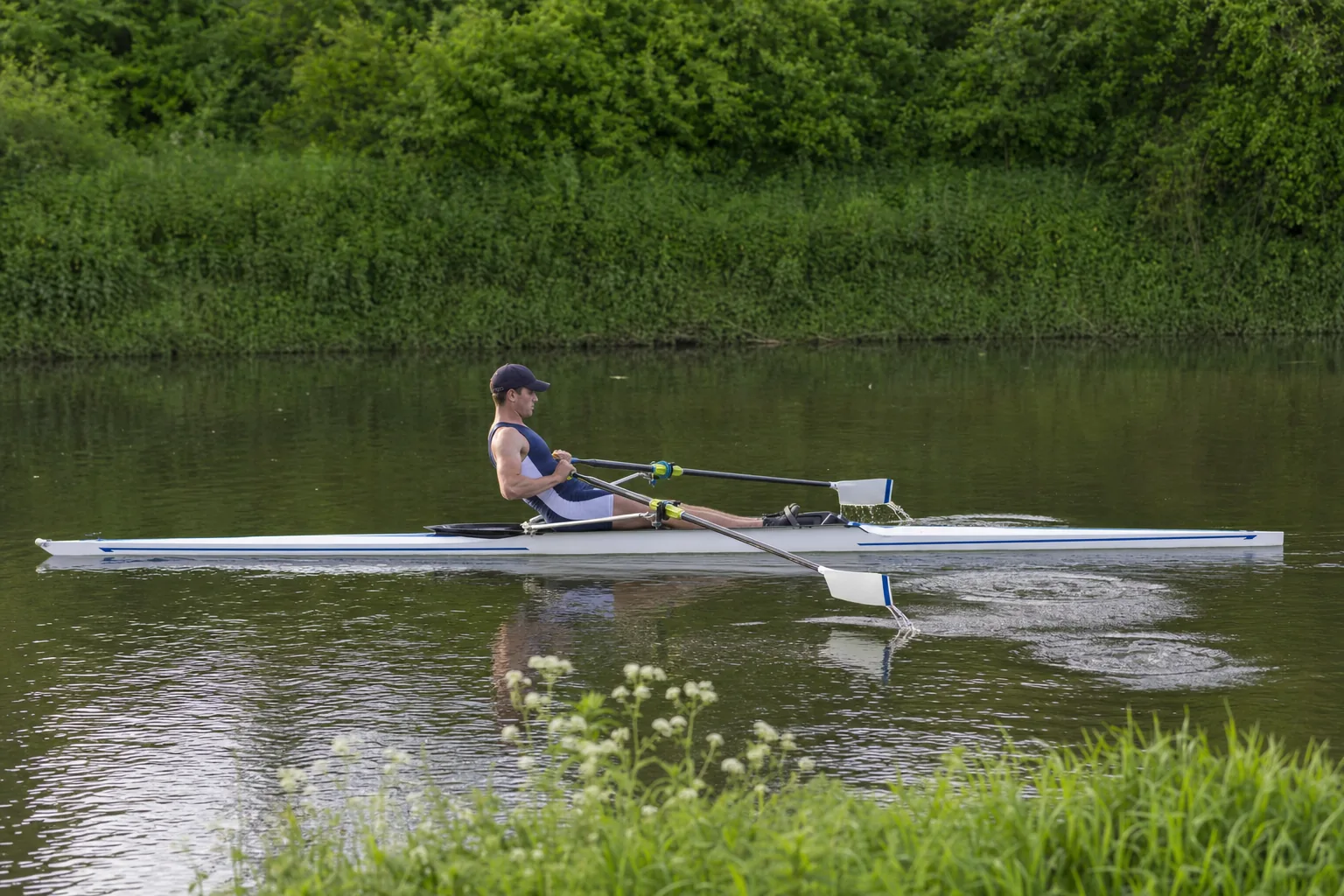 A single sculler at the finish, legs flat, body leaning back and hands drawn in, with both blades lifting clear of the water and puddles left behind
